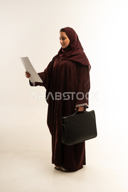 قراءة الملاحظات والمعلومات، الأناقة والاهتمام بالمظهر الخارجي، مفهوم إدارة الأعمال والمشاريع، بورتريه لامراة عربية خليجية عمانية محجبة ترتدي عباءة بنيه تحمل بيدها حقيبه سوداء، سيده سعودية تنظر الى مجموعه من الاوراق بايماءات السرور، الجسم بالطول الكامل، خلفية بيضاء