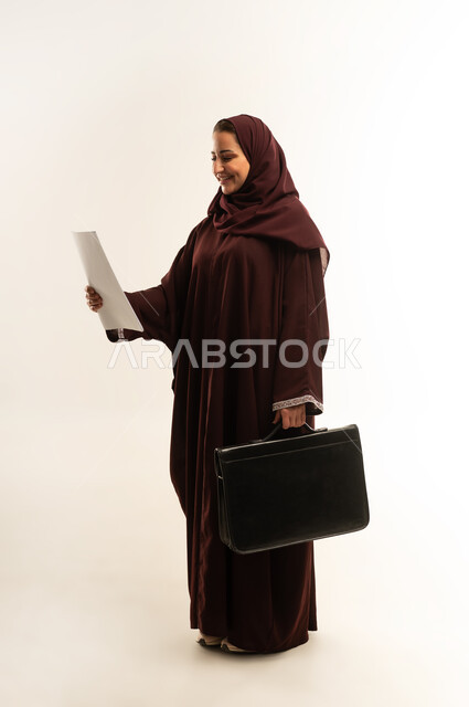 Elegance and interest in outward appearance, the concept of business and project management, portrait of a veiled Omani Gulf Arab woman wearing a brown abaya, carrying a black bag in her hand, a Saudi woman looking at a group of papers with gestures of pleasure, full body, white background