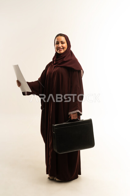 مفهوم إدارة الأعمال والمشاريع، تعابير السعادة والسرور، بورتريه لامراة عربية خليجية عمانية محجبة ترتدي عباءة بنيه تحمل بيدها مجموعه من الاوراق و حقيبه سوداء، سيده سعودية تنظر الى الكاميرا بايماءات الثقة بالنفس، الجسم بالطول الكامل، خلفية بيضاء