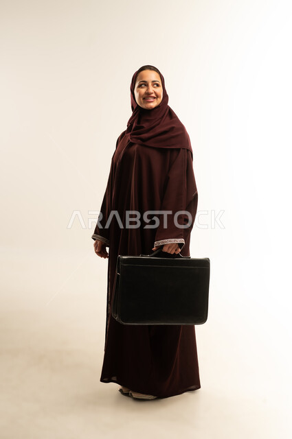 الأناقة والاهتمام بالمظهر الخارجي، مفهوم إدارة الأعمال والمشاريع، سيده سعودية تستعد للذهاب إلى العمل، بورتريه لامراه عربيه خليجيه عمانيه مبتسمه ترتدي العباءه تحمل بيديها حقيبة يد و تقوم بالنظر الى شيئ ما، الجسم بالطول الكامل، خلفيه بيضاء