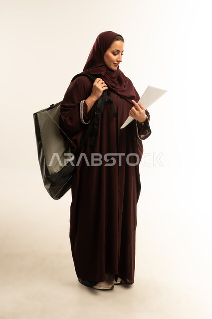 الجامعات والكليات الأكاديمية في سلطنة عمان، التحضير للذهاب إلى الجامعة، بورتريه لطالبة جامعية عربية عمانية خليجية محجبة مبتسمه تحمل حقيبة على كتفها ، سيده سعوديه تحمل بيدها الأوراق, الجسم بالطول الكامل، خلفية بيضاء