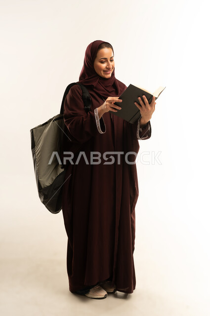 University education, reviewing daily lessons, the concept of diligence and excellence, portrait of an Arab Gulf Omani university student wearing an abaya, carrying a bag on her back and holding a book studying lectures, a Saudi woman reading university books, full body, white background