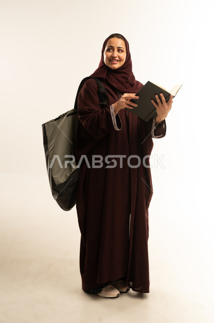 مراجعة الدروس اليومية، مفهوم الاجتهاد والتفوق, التعليم الجامعي, بورتريه لطالبه جامعيه عربيه خليجيه عمانيه ترتدي العباءه تحمل حقيبة على ظهرها وبيدها كتاب تقوم بدراسة المحاضرات, سيده سعوديه تنظر الى شيء ما ، الجسم بالطول الكامل, خلفية بيضاء