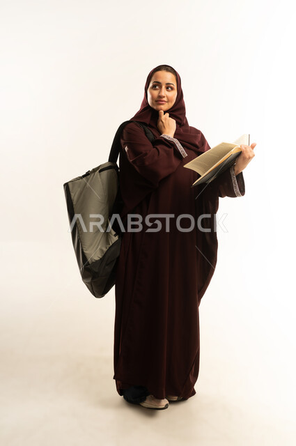 مفهوم الاجتهاد والتفوق, مراجعة الدروس اليومية, التعليم الجامعي, بورتريه لطالبه جامعيه عربيه خليجيه عمانيه ترتدي العباءه تحمل حقيبة على ظهرها وتمسك بيدها كتاب تقوم بدراسة المحاضرات, سيده سعوديه تفكر بشي ما ، الجسم بالطول الكامل, خلفية بيضاء