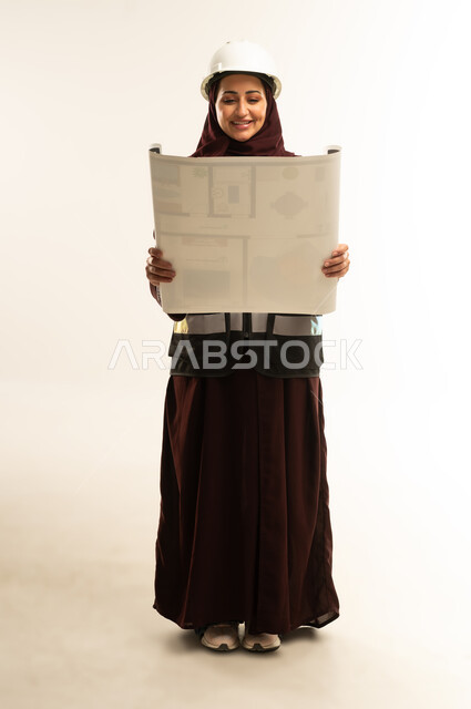 Reviewing engineering plans, supervising work progress, portrait of a veiled Gulf Arab Omani female engineer wearing a safety helmet and work jacket, holding a construction plan in her hand, Saudi women working in the engineering sector, full body, white background