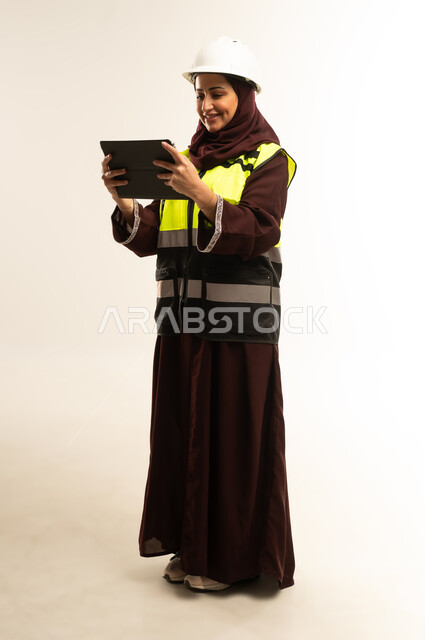 Expressions of focus and preoccupation, completing tasks remotely, managing engineering projects, working in the engineering sector, portrait of an Arab Gulf Omani female engineer wearing a protective jacket and helmet working on a tablet, a young Saudi woman studying the basics of the project, full body, white background
