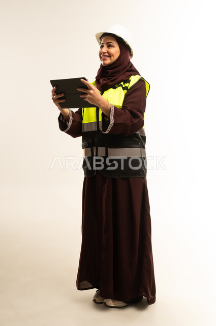 العمل في القطاع الهندسي، انجاز المهام عن بعد، إدارة المشاريع الهندسية, بورتريه لمهندسة عربية خليجية عمانية ترتدي سترة وخوذة الحماية تعمل على جهاز التابلت, شابة سعودية تقوم بدراسة أساسيات المشروع، الجسم بالطول الكامل، خلفية بيضاء