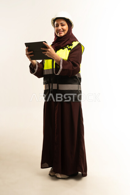 Completing tasks remotely, managing engineering projects, working in the engineering sector, portrait of an Arab Gulf Omani female engineer wearing a protective jacket and helmet working on a tablet, a young Saudi woman studying the basics of the project, full body, white background