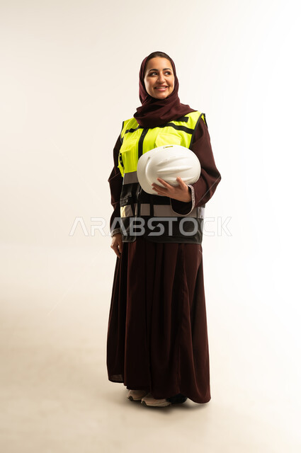 Engineering and construction concept, female engineering professions and jobs, portrait of a smiling Gulf Arab Omani female engineer wearing hijab and a protective vest working in the engineering field, a Saudi woman standing straight holding a helmet in her hands and looking at something, full body image, white background
