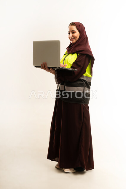 مهن ووظائف نسائية، مشروع بناء، مفهوم الهندسه المعماريه، بورتريه لمهندسة معمارية عربية عمانيه خليجية مبتسمه تحمل بيدها جهاز اللابتوب المحمول ، مهندسة إنشاءات محجبة ترتدي بدلة السلامة في العمل, الجسم بالطول الكامل، خلفية بيضاء