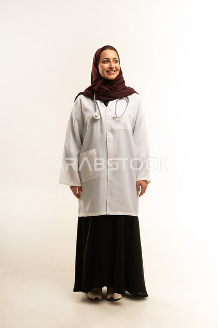 Providing medical services and examinations to patients, a smiling Saudi woman standing straight and looking at the camera with confidence, working in the health sector, a picture of a smiling veiled Omani Gulf Arab female doctor wearing a white medical coat, gestures of happiness and pleasure