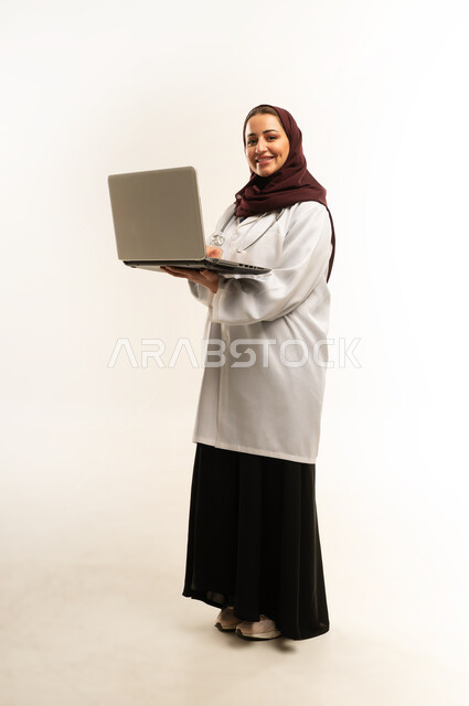 مفهوم استخدام أجهزة تقنيه حديثه، العمل في القطاع الصحي، بورتريه لطبيبة عربية خليجية عمانيه مبتسمه محجبة ترتدي المعطف الطبي ، سيده سعودية تحمل بيدها الحاسوب المحمول,الجسم بالطول الكامل، خلفية بيضاء