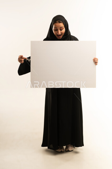 لوحات توضيحية ووسائل تعليمية، عرض و اعلان تسويقي, بورتريه لإمرأة عربية خليجية عمانية محجبة ترتدي العباءة السوداء تمسك بيديها لوحة بيضاء مستطيلة فارغة، شابة سعودية تنظر بايماءات الدهشه و الاعجاب, موك اب مستطيل أبيض, الجسم بالطول الكامل, خلفية بيضاء