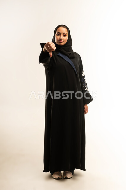 القلق والفشل في تحقيق أمر ما، مفهوم التقييم السلبي والانزعاج، بورتريه لامراه عربيه خليجيه عمانية تنظر الى الكاميرا و تشير بإصبع الابهام الى الاسفل بإيماءات الانزعاج، سيده سعودية تشعر بالرفض وعدم القبول والرضا، الجسم بالطول الكامل، خلفية بيضاء