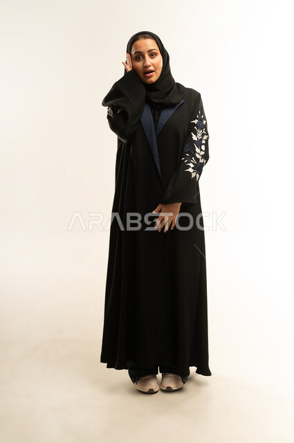 Looking at the camera with expressions of surprise and astonishment, a Saudi woman wondering what happened, a portrait of a Saudi Omani Gulf Arab woman wearing a black abaya, putting her hands on her face with gestures indicating shock and astonishment, full body, white background
