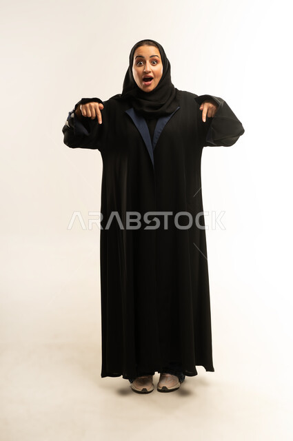التعجب والتفاجؤ من مشاهدة شيء ما، سيده سعودية تنظر إلى الكاميرا بتعابير الاستغراب والاندهاش, بورتريه لامرأة عربية خليجية عمانية محجبة ترتدي عباءة سوداء تشاور بيديها الى شيء ما بإيماءات الذهول والصدمة، صورة الجسم بالطول الكامل، خلفية بيضاء