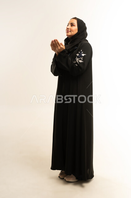 التقرب إلى الله تعالى والرغبة في تحقيق الأمنيات، الترجي والتذلل لرب العالمين, الدعاء والتضرع والتوسل بإيماءات الخشوع, سيدة سعودية ترتدي العباءة السوداء تنظر إلى الأعلى, بورتريه لإمرأة عربية خليجية عمانيه ترفع يديها باستعطاف, الجسم بالطول الكامل, خلفية بيضاء