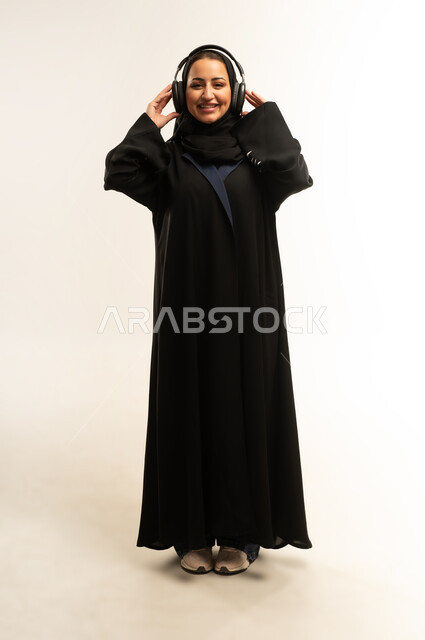 التسلية والترفيه بإستخدام تقنية الهيدفون، بورتريه لامراه عربيه خليجيه عمانيه  مبتسمه ترتدي العباءه السوداء تستمتع بسماع الأغاني والموسيقى والصوتيات،سيده سعوديه ترتدي سماعات الرأس الصوتية، الجسم بالطول الكامل، خلفية بيضاء