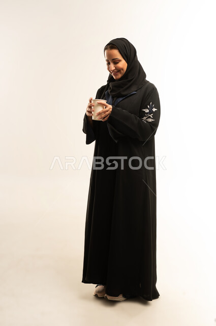 قضاء وقت ممتع و هادئ، الحيوية والنشاط عند شرب الكافيين، بورترية لامراه عربيه خليجيه عمانيه مبتسمه ترتدي العباءه السوداء تقوم بالاستمتاع بتناول المشروبات الساخن، سيده سعوديه تحمل بيديها كوب ورقي، الجسم بالطول الكامل، خلفية بيضاء