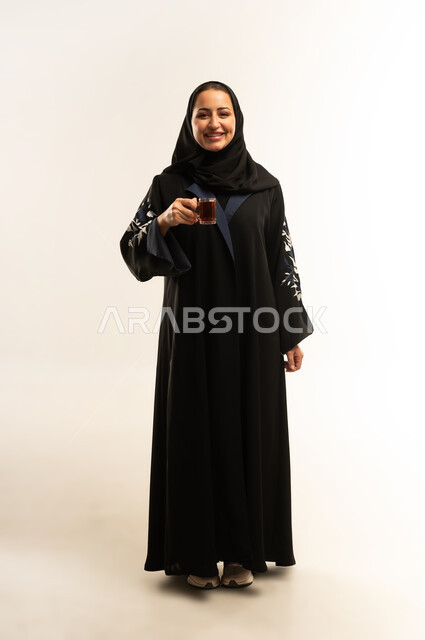 احتساء المشروبات الساخنه،  سيده سعوديه مبتسمه تتذوق المشروب المفضل، بورتريه لامراه عربيه خليجيه عمانيه ترتدي العباءه السوداء تمسك بيديها كوب من الشاي , تعابير السعاده و السرور، قضاء وقت ممتع و هادئ، الجسم بالطول الكامل، خلفية بيضاء