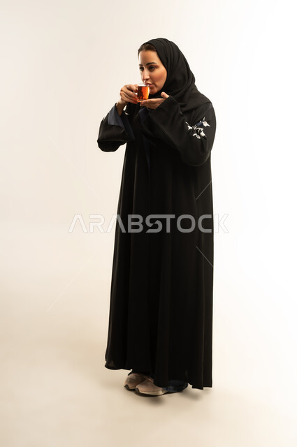 قضاء وقت ممتع و هادئ، الاستمتاع بتناول المشروبات الساخنه الشهيه ، سيده سعوديه مبتسمه تتذوق المشروب المفضل، بورتريه لامراه عربيه خليجيه عمانيه ترتدي العباءه السوداء تقوم بتناول كوب من الشاي , تعابير السعاده و السرور، الجسم بالطول الكامل، خلفية بيضاء