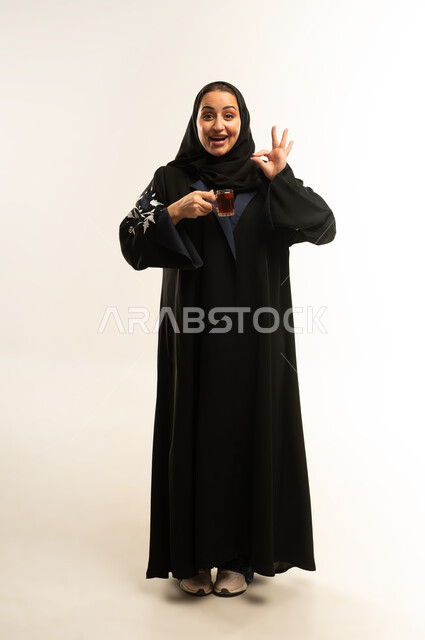 تعابير الرضا والموافقة, مفهوم القبول والإعجاب، بورتريه لامراة عربيه خليجيه عمانيه ترتدي العباءه السوداء تقوم بشرب فنجان من الشاي الساخن, سيده سعوديه ترفع يديها نحو الأعلى بعلامة الجودة, الجسم بالطول الكامل، خلفية بيضاء