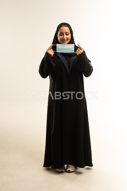 مفهوم الوعي الصحي, سيده سعوديه تقوم بالإلتزام بالإجراءات الوقايه والسلامة للحمايه من العدوى بكوفيد-١٩، بورتريه لامرأة عربية خليجية عمانية محجبة ترتدي عباءه سوداء تضع الكمامة لتجنب تفشي فايروس كورونا، الجسم بالطول الكامل، خلفية بيضاء