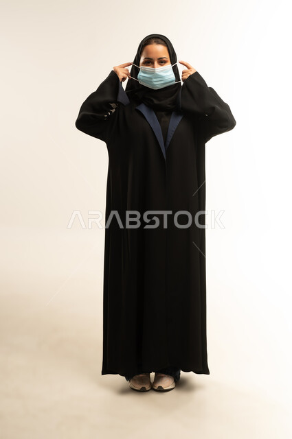 الحفاظ على السلامة من الأمراض والفيروسات, سيده سعوديه تقوم بالإلتزام بالإجراءات الوقايه والسلامة للحمايه من العدوى بكوفيد-١٩، بورتريه لامرأة عربية خليجية عمانية محجبة ترتدي عباءه سوداء تضع الكمامة لتجنب تفشي فايروس كورونا، الجسم بالطول الكامل، خلفية بيضاء