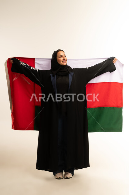 Celebrating Oman's National Day, participating in national holidays and occasions, pride in Omani symbols and flag, portrait of a smiling veiled Omani Gulf Arab woman wearing a black abaya, carrying the flag of the Sultanate of Oman on her back and looking at something, full body, white background