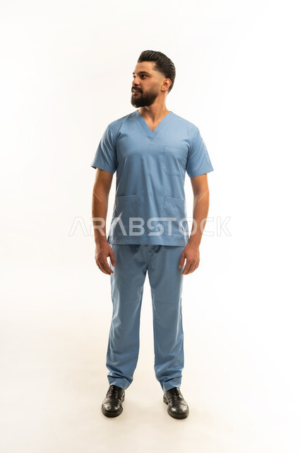 الاستعداد لتقديم الخدمات الطبية للمرضى, مفهوم الطب والرعاية الصحية، ممرض سعودي يعمل بالقطاع الطبي، الوقوف باستعداد والنظر إلى شيء ما بإيماءات السرور, بورتريه لشاب عربي خليجي عماني مبتسم يرتدي يونيفورم العمليقف باستقامة, الجسم بالطول الكامل، خلفية بيضاء