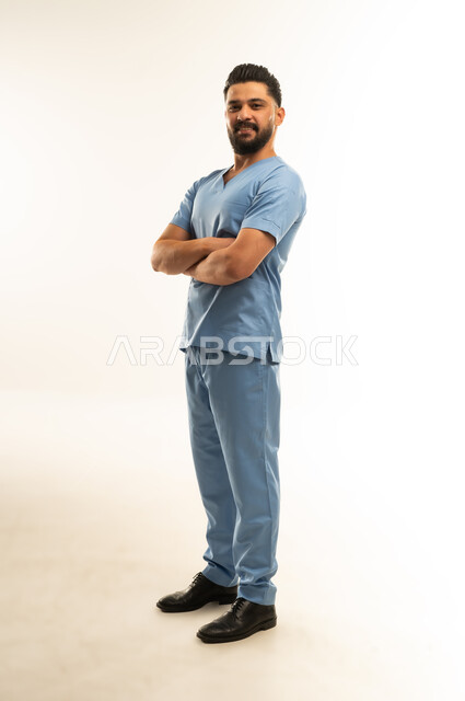 الوقوف بأيدي متقاطعة بتعابير الثقة بالنفس, مفهوم الطب والرعاية الصحية، الاستعداد لتقديم الخدمات الطبية للمرضى,  ممرض سعودي يعمل بالقطاع الطبي، بورتريه لشاب عربي خليجي عماني مبتسم يرتدي يونيفورم العمليقف باستقامة, الجسم بالطول الكامل، خلفية بيضاء