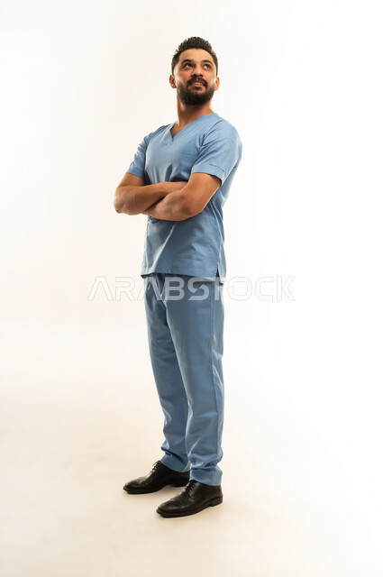 الوقوف بأيدي متقاطعة بتعابير الثقة بالنفس, مفهوم الطب والرعاية الصحية، الاستعداد لتقديم الخدمات الطبية للمرضى,  ممرض سعودي يعمل بالقطاع الطبي، بورتريه لشاب عربي خليجي عماني مبتسم يرتدي يونيفورم العمليقف باستقامة, الجسم بالطول الكامل، خلفية بيضاء