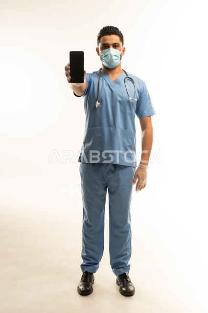 عرض شاشة سوداء فارغة، استخدام التقنية الحديثه في المجال الطبي، ممرض سعودي يمسك بيده الهاتف المحمول, بورتريه لشاب عربي خليجي عماني يرتدي يونيفورم العمل والكمامة يعمل في المجال الصحي، الجسم بالطول الكامل، خلفية بيضاء