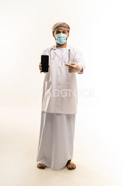 عرض شاشة سوداء فارغة عبر الهاتف المحمول, استخدام التقنية الحديثه في المجال الطبي، تقديم الخدمات العلاجية, بورتريه لطبيب عربي خليجي عماني يرتدي الدشداشة والعمامة والمعطف الطبي يضع السماعة الطبية يعمل في  العمل في القطاع الصحي، الجسم بالطول الكامل، خلفية بيضاء