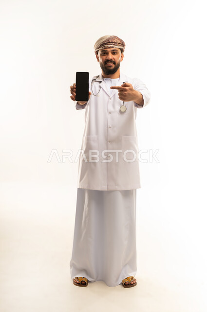 عرض شاشة سوداء فارغة عبر الهاتف المحمول, استخدام التقنية الحديثه في المجال الطبي، تقديم الخدمات العلاجية, بورتريه لطبيب عربي خليجي عماني يرتدي الدشداشة والعمامة والمعطف الطبي يضع السماعة الطبية يعمل في  العمل في القطاع الصحي، الجسم بالطول الكامل، خلفية بيضاء