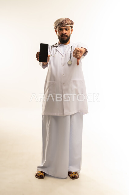 خفض اصبع الابهام بتعابير الغضب وعدم الاعجاب، عرض شاشة سوداء فارغة عبر الهاتف المحمول, استخدام التقنية الحديثه في الطب، بورتريه لطبيب عربي خليجي عماني يرتدي الدشداشة والعمامة والمعطف الطبي يضع السماعة الطبية يعمل في القطاع الصحي، الجسم بالطول الكامل، خلفية بيضاء