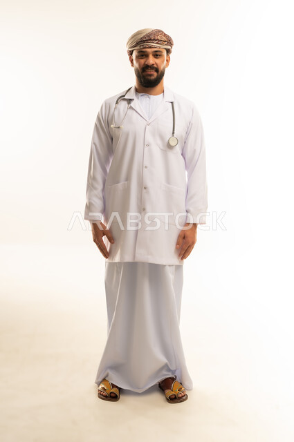 Working in the health sector in the Sultanate of Oman, medical examination and providing treatment services to patients, portrait of an Arab Gulf Omani doctor wearing a dishdasha, turban and medical coat, wearing a stethoscope, standing straight, looking at the camera with self-confident gestures, full body, white background