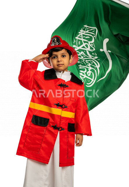 بورتريه لفتى عربي خليجي سعودي أنيق ، يرتدي زي رجل الإطفاء ، يلقي التحية لوطنه الحبيب المملكة العربية السعودية ، خلفه علم المملكة ، اليوم الوطني السعودي ، مفهوم مهنة الدفاع المدني ، المهنة المستقبلية ، خلفية بيضاء ، يرتدي خوذة وسترة الحماية الخاصة بالعمل