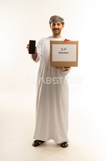 عرض شاشة سوداء فارغة عبر الهاتف المحمول، المساعدة والتبرع للسلة الخيرية، بورتريه لرجل عربي خليجي عماني يرتدي الدشداشة والعمامة يحمل صندوق التبرع، جمعية ومبادرة إنسانية في سلطنة عمان، توفير الطعام للفقراء، الجسم بالطول الكامل، خلفية بيضاء