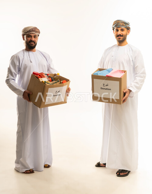 Providing aid to the needy, providing essential supplies to the poor, a humanitarian initiative in the Sultanate of Oman, the concept of assistance, donation and charity, a portrait of two Omani Gulf Arab men wearing dishdashas and turbans preparing donation boxes, full body, white background