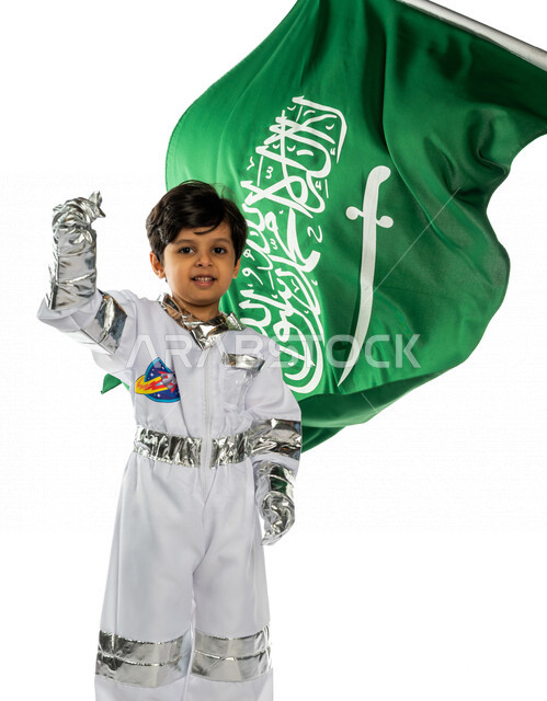 بورتريه لفتى عربي خليجي سعودي مبتسم ، يرتدي زي رائد الفضاء ، يقوم بإيماءات يد مختلفة تدل على حب الوطن ، خلفه علم المملكة العربية السعودية ، ذكرى اليوم الوطني السعودي ، يحلم بأن يصبح رائد فضاء ، حلم الوصول للفضاء والقمر ، خلفية بيضاء
