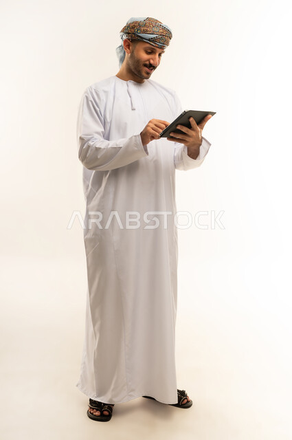 Using modern technology and techniques, browsing social networking sites via tablet, chatting with family and friends, portrait of an Arab Gulf Omani man wearing a dishdasha and turban holding a tablet in his hand, full body length, white background
