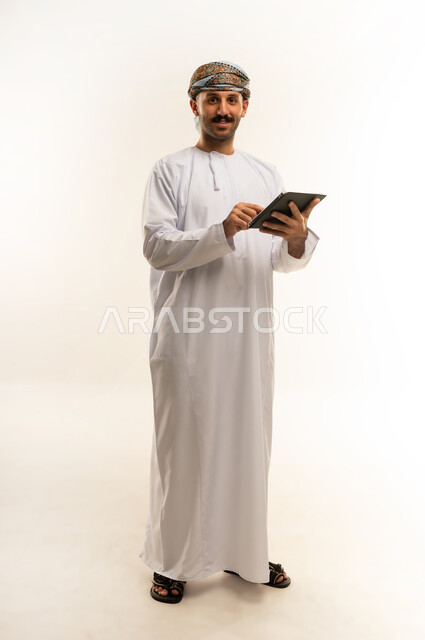 استعمال التكنولوجيا والتقنية الحديثة, تصفح مواقع التواصل الاجتماعي عبر الجهاز اللوحي، المحادثة مع الأهل والأصدقاء، بورتريه لرجل عربي خليجي عماني يرتدي الدشداشة والعمامة يحمل بيده جهاز التابلت، الطول الكامل للجسم، خلفية بيضاء