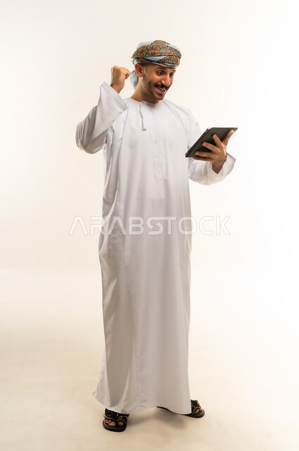 تعابير الفوز والانتصار، استعمال التكنولوجيا والتقنية الحديثة, تصفح مواقع التواصل الاجتماعي عبر الجهاز اللوحي، المحادثة مع الأهل والأصدقاء، بورتريه لرجل عربي خليجي عماني يرتدي الدشداشة والعمامة يحمل بيده جهاز التابلت، الطول الكامل للجسم، خلفية بيضاء