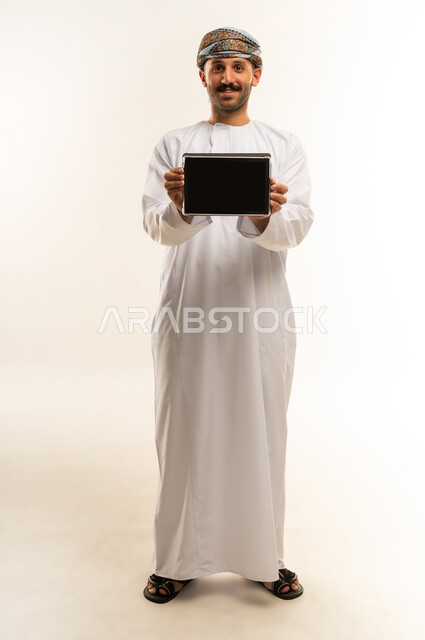 Blank black screen, using modern technology, browsing social media on a tablet, chatting with family and friends, portrait of an Arab Gulf Omani man wearing a dishdasha and turban holding a tablet, full body length, white background