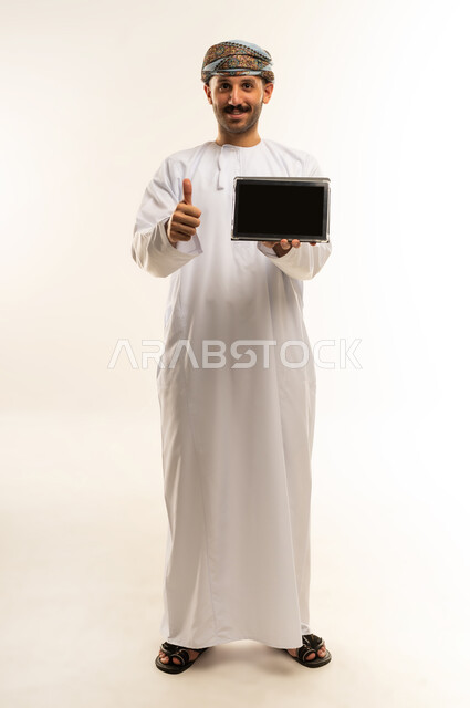 تعابير الاعجاب والقبول والرضا، عرض شاشة سوداء فارغة، استعمال التكنولوجيا والتقنية الحديثة, تصفح مواقع التواصل الاجتماعي عبر الجهاز اللوحي، بورتريه لرجل عربي خليجي عماني يرتدي الدشداشة والعمامة يحمل بيده جهاز التابلت، الطول الكامل للجسم، خلفية بيضاء