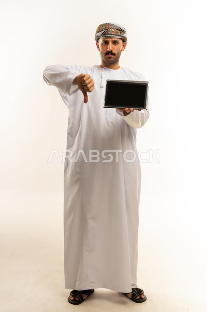 تعابير عدم الاعجاب والرفض، عرض شاشة سوداء فارغة، استعمال التكنولوجيا والتقنية الحديثة, تصفح مواقع التواصل الاجتماعي عبر الجهاز اللوحي، بورتريه لرجل عربي خليجي عماني يرتدي الدشداشة والعمامة يحمل بيده جهاز التابلت، الطول الكامل للجسم، خلفية بيضاء