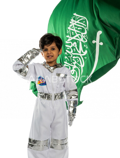 بورتريه لفتى عربي خليجي سعودي مبتسم ، يرتدي زي رائد الفضاء ، يلقي التحية لوطنه الحبيب المملكة العربية السعودية ، خلفه علم المملكة ، ذكرى اليوم الوطني السعودي ،الاحتفال بالعيد الوطني السعودي ، يحلم بأن يصبح رائد فضاء ، خلفية بيضاء