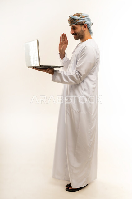 Using modern technology, the concept of diligence and completing required tasks, a portrait of a young Arab Gulf Omani man wearing a dishdasha and turban, standing and holding a laptop in his hand, browsing social networking sites, integrating technology into daily life, a full-length body image, white background