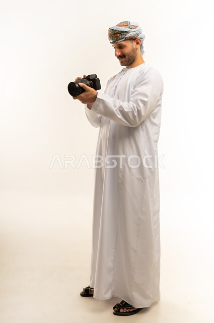 Practicing the hobby of photography, a professional photographer taking a souvenir photo, using high-resolution digital cameras, a close-up portrait of an Arab Gulf Omani man wearing a dishdasha and turban, holding a camera in his hand, taking photographs with gestures of concentration and contemplation, full-length body, white background